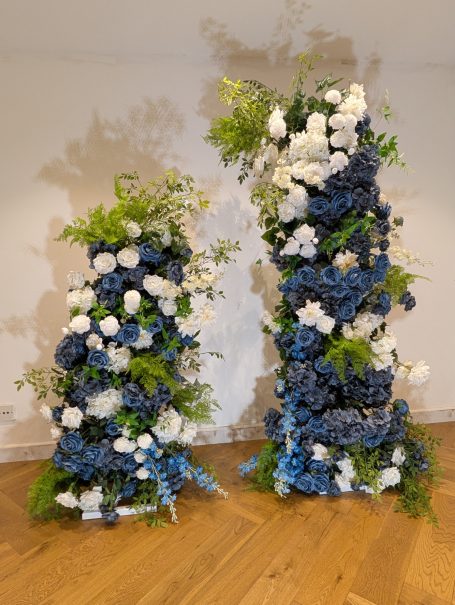 Broken Arch "Isabella" Two floral arrangements featuring blue and white flowers, displayed on wooden flooring.